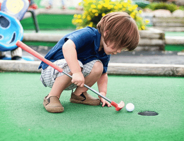 little boy with a golf club looking at a golf ball on a putting green