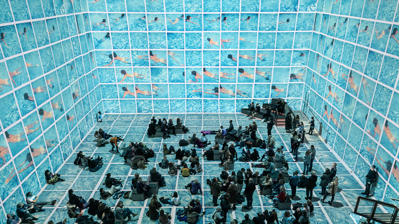 Families relax on a floor of rippling blue water as Hockney's swimmers fill every wall at the Bigger & Closer immersive show