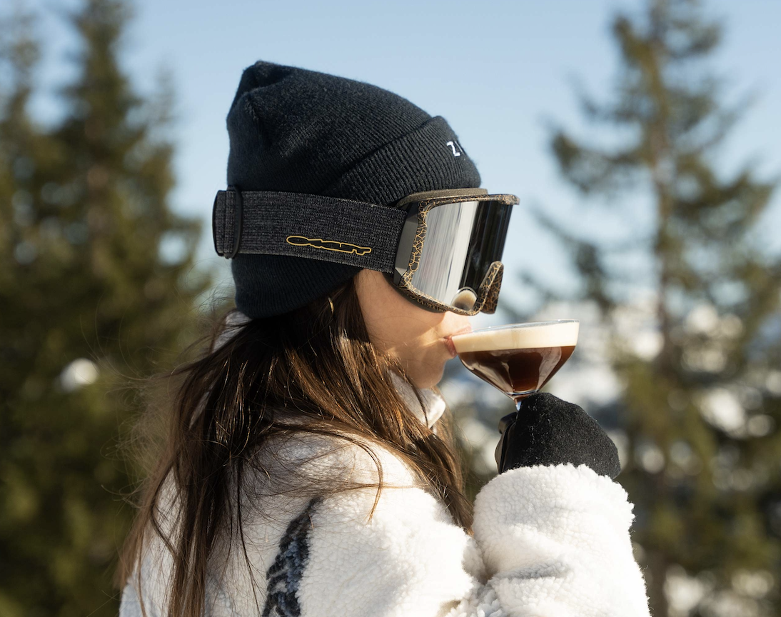 Skier sipping an après cocktail at Zuma in the sunshine, goggles on and snowy pine trees behind