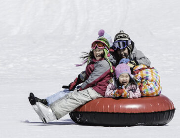 Four laughing kids pile onto one snow tube and slide down a sunny slope at Hanazono Tube Park, Niseko.
