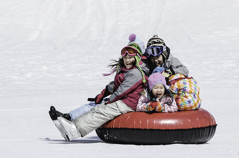 Four laughing kids pile onto one snow tube and slide down a sunny slope at Hanazono Tube Park, Niseko.